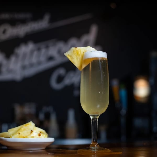 A pineapple mimosa with a pineapple wedge garnish and a side of pineapple chunks on a bar.