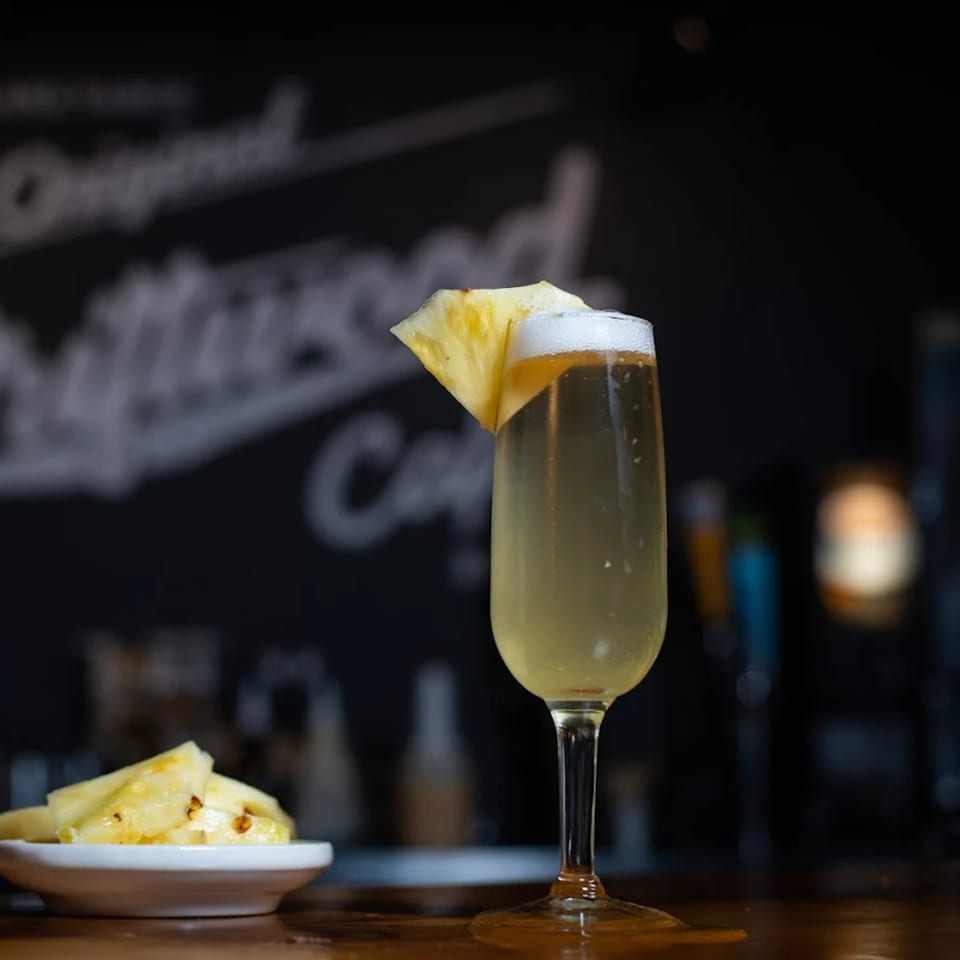 A pineapple mimosa with a pineapple wedge garnish and a side of pineapple chunks on a bar.
