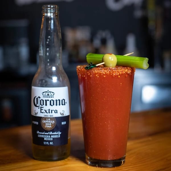 A Corona Extra bottle next to a Bloody Mary cocktail garnished with celery and an olive.