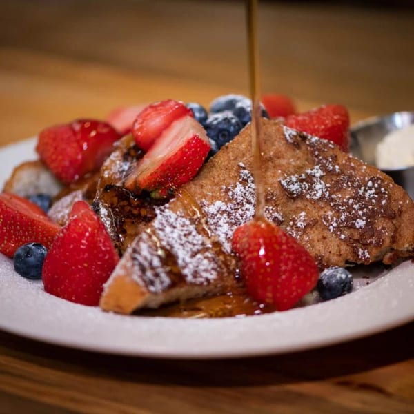 Syrup pouring over French toast with powdered sugar, strawberries, and blueberries on a white plate.