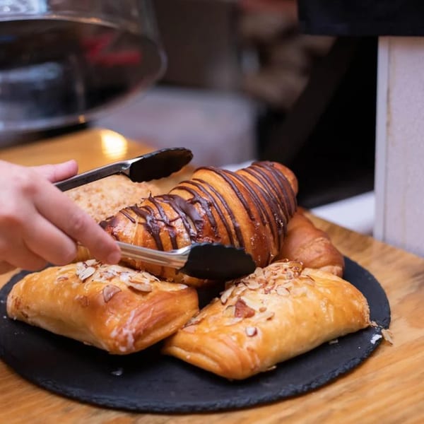 A hand uses tongs to pick up a chocolate drizzled croissant from a platter of pastries.