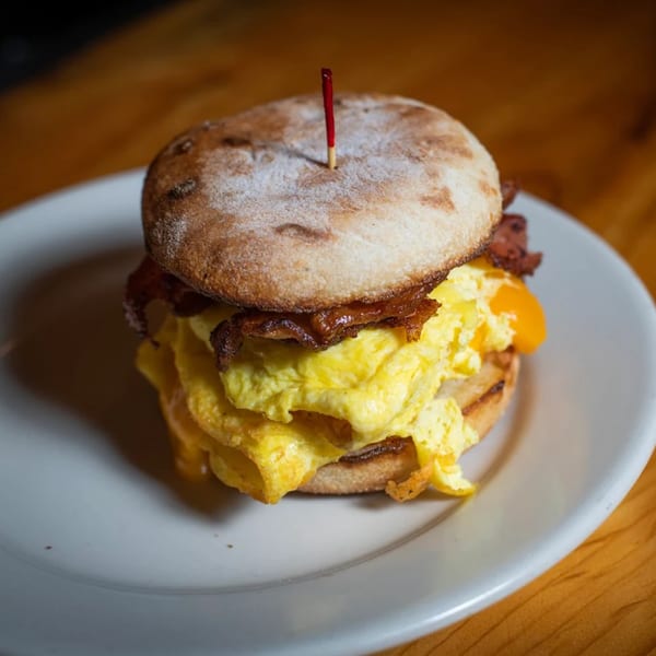 A breakfast sandwich with scrambled eggs, bacon, and cheese on an English muffin, served on a white plate.