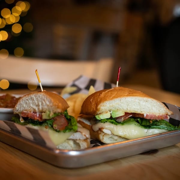Two gourmet sandwiches with cheese, avocado, and bacon on a metal tray at Driftwood Cafe.