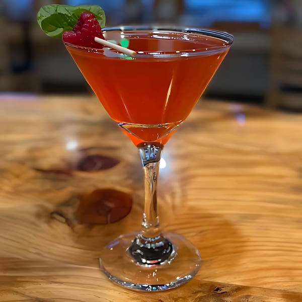 A red cocktail in a martini glass, garnished with raspberries and a basil leaf on a wooden table.